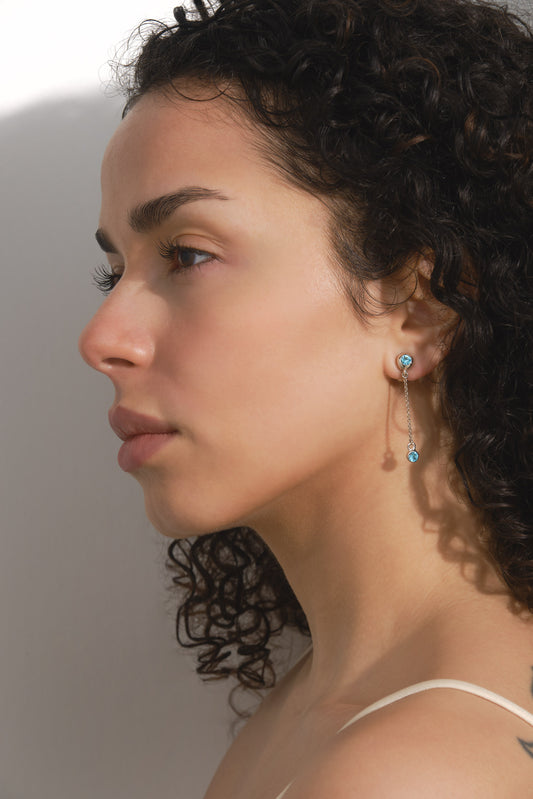 A close-up side profile of a woman with curly dark hair wearing a delicate drop earring with a silver-toned circular stud and a small dangling turquoise-blue bead.