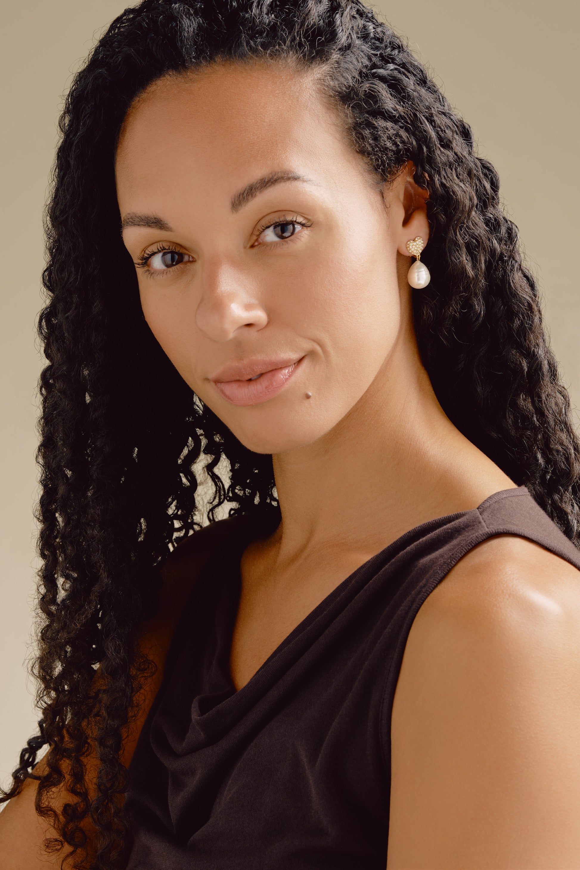 Woman with long, curly hair wearing a black top against a neutral background wearing gold heart and pearl earrings