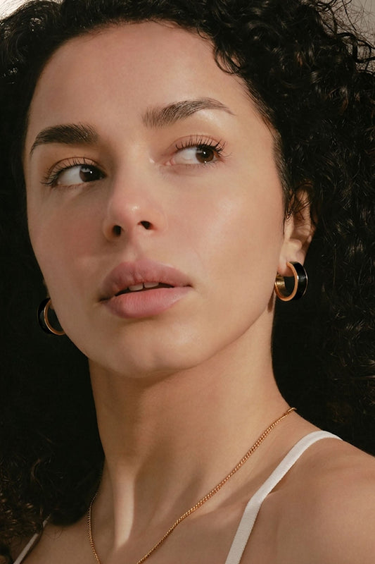 A close-up lifestyle shot of a woman with curly dark hair wearing a delicate gold necklace with a small horizontal bar pendant and matching gold hoop earrings.