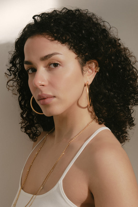 A close-up portrait of a woman with curly dark hair wearing large gold hoop earrings, a delicate gold chain necklace, and a gold ear cuff, paired with a white camisole top against a warm neutral background.