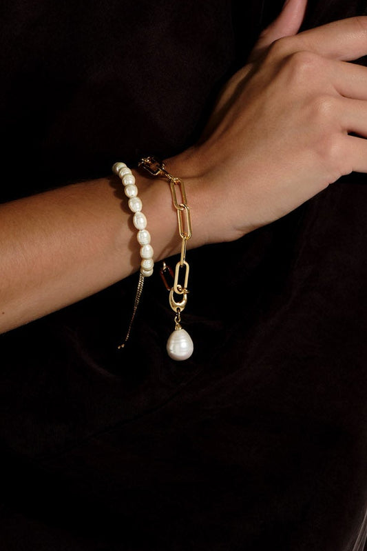 A woman with long brown hair models a gold chain necklace with a white pearl pendant and matching pearl-and-gold bracelets against a white wall.