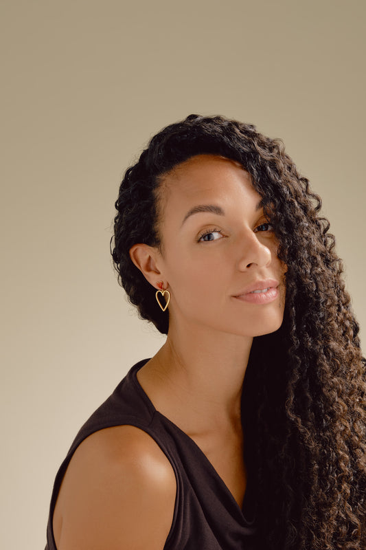 Woman with long, curly hair wearing a black sleeveless top against a beige background wearing red heart earrings