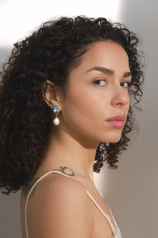 A close-up portrait of a woman with curly dark hair wearing a dangling earring with a turquoise-blue top and a white pearl drop, photographed against a soft light gray background.