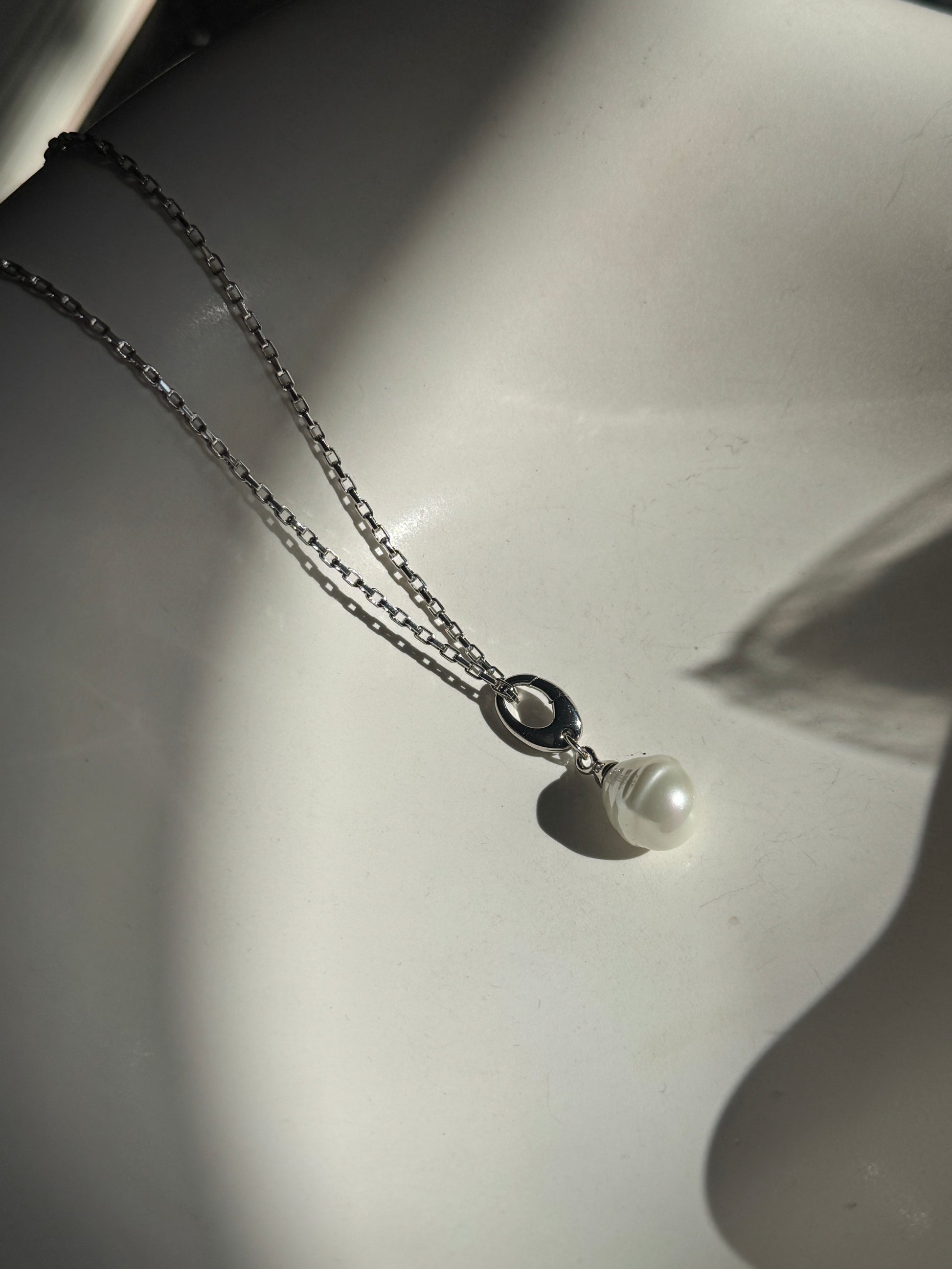 A silver chain necklace with a small silver ring connector and a single white pearl pendant, displayed on a white surface with dramatic shadows.