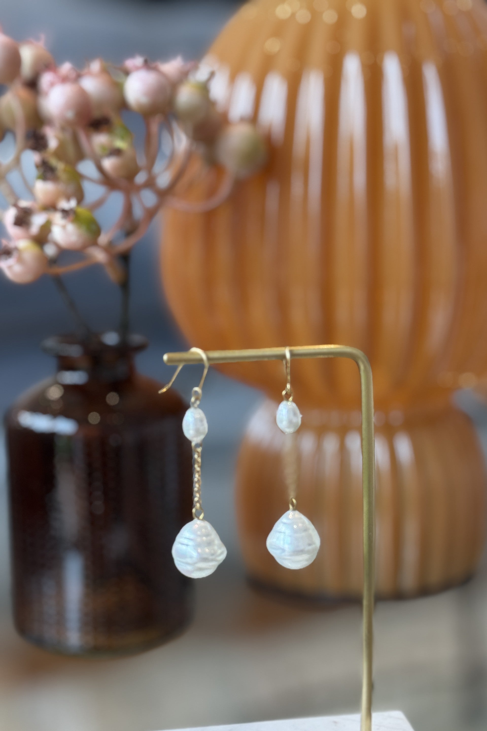 Pearl earrings on a gold stand with a blurred background featuring a vase and decorative item.