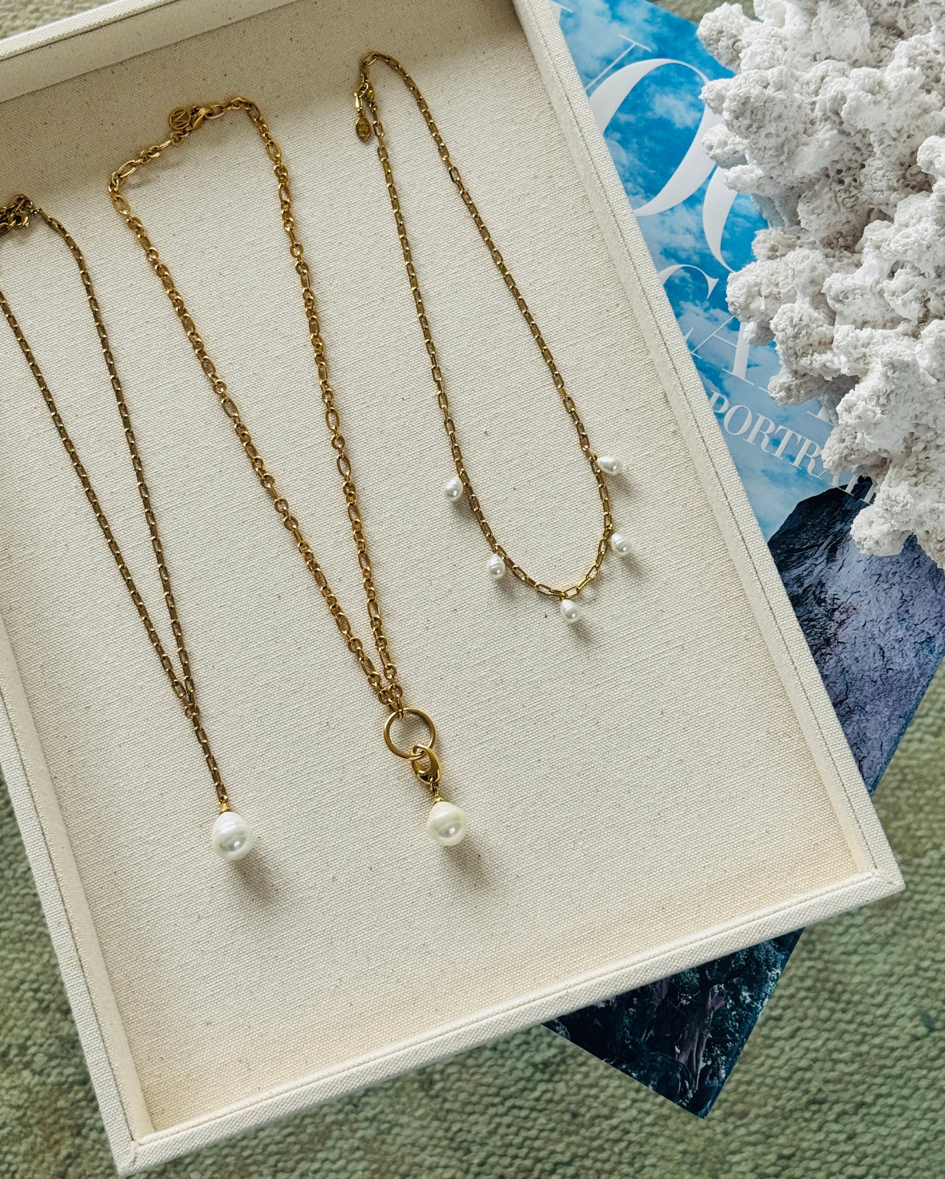 Two gold necklaces with pearl pendants on a textured surface.