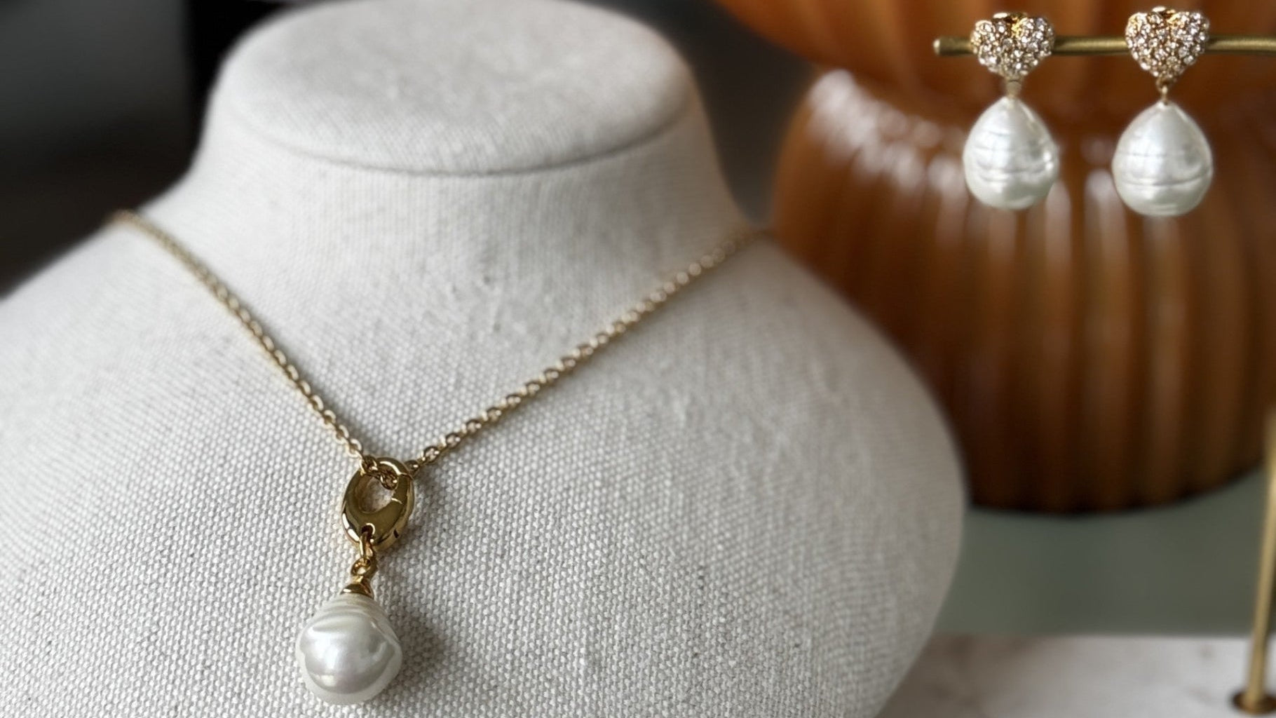 Gold necklace with a pearl pendant on a mannequin bust, pearl earrings on a stand, and decorative vase in the background.