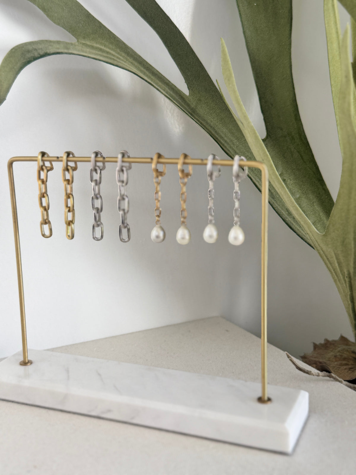A gold metal jewelry display stand with a white marble base, holding several earrings including gold and silver chain links and pearl drops, styled with a green plant in the background.