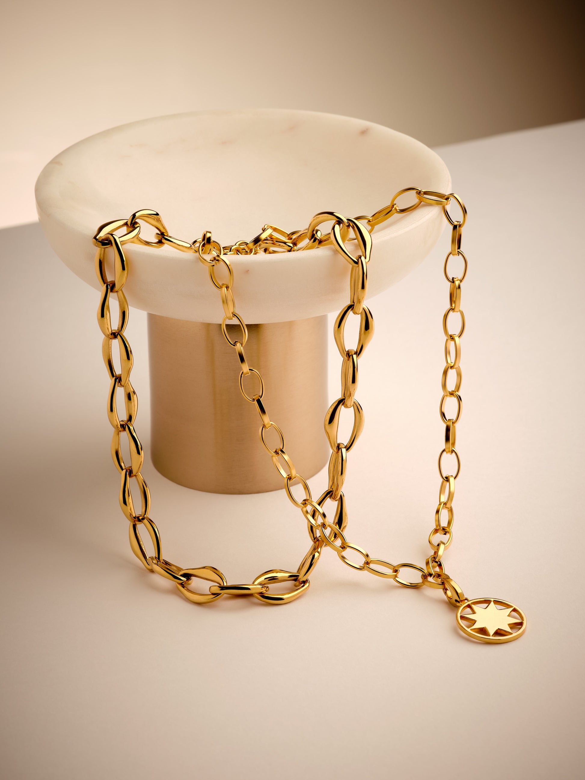 Gold chain necklace with a star pendant on a white pedestal against a beige background