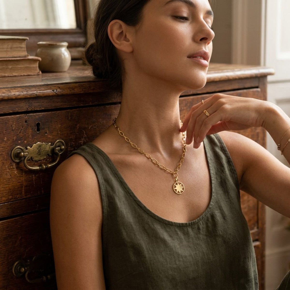 A woman wearing a delicate gold chain necklace with a circular sunburst pendant, leaning against a rustic wooden dresser.
