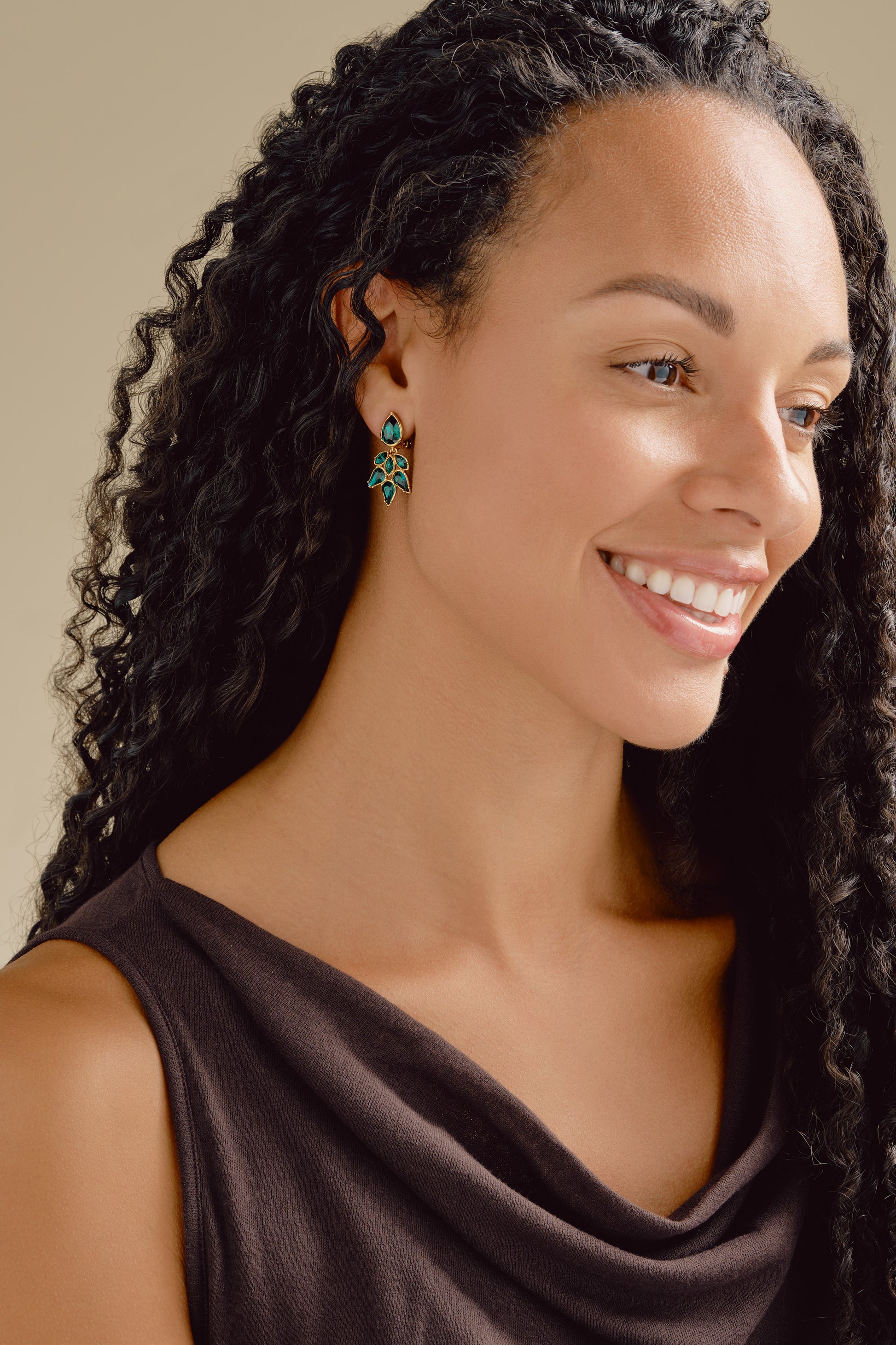 Woman with braided hair wearing a dark brown top against a neutral background wearing emerald earrings
