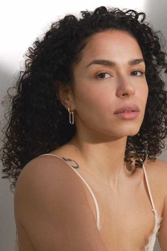 Woman with curly hair wearing a sheer top against a neutral background with gold and silver earrings