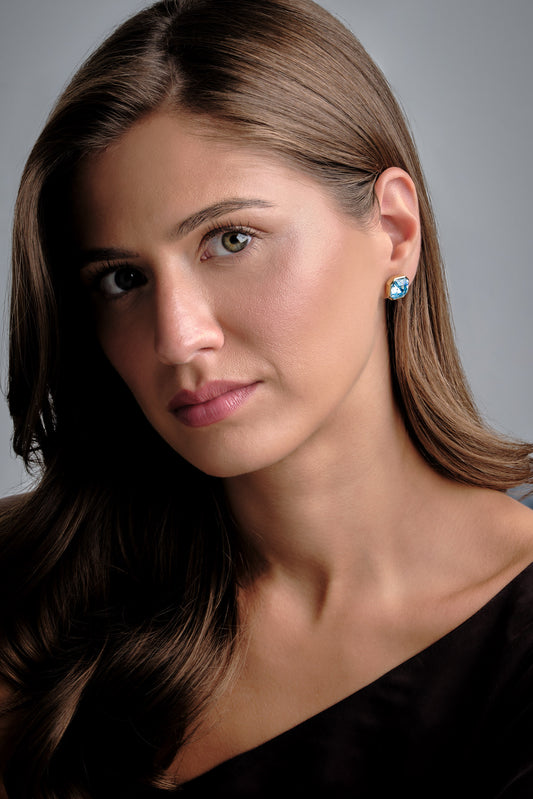 A close-up portrait of a woman with long brown hair wearing a small light blue and white stud earring, set against a plain gray studio background.