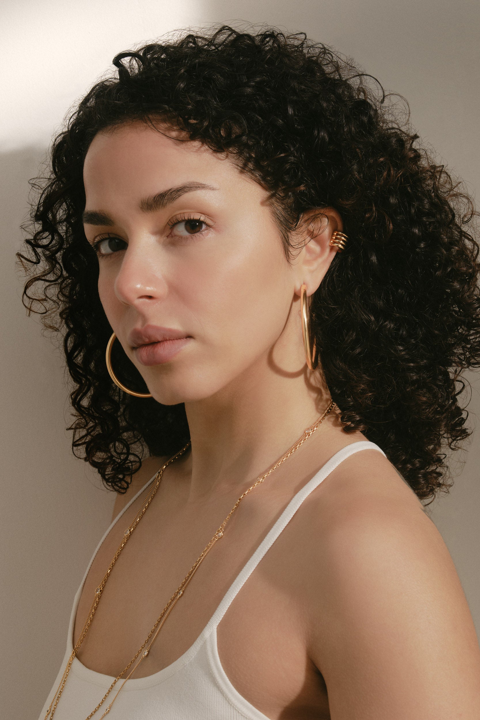 A close-up portrait of a woman with curly dark hair wearing large gold hoop earrings, a delicate gold chain necklace, and a gold ear cuff, paired with a white camisole top against a warm neutral background.