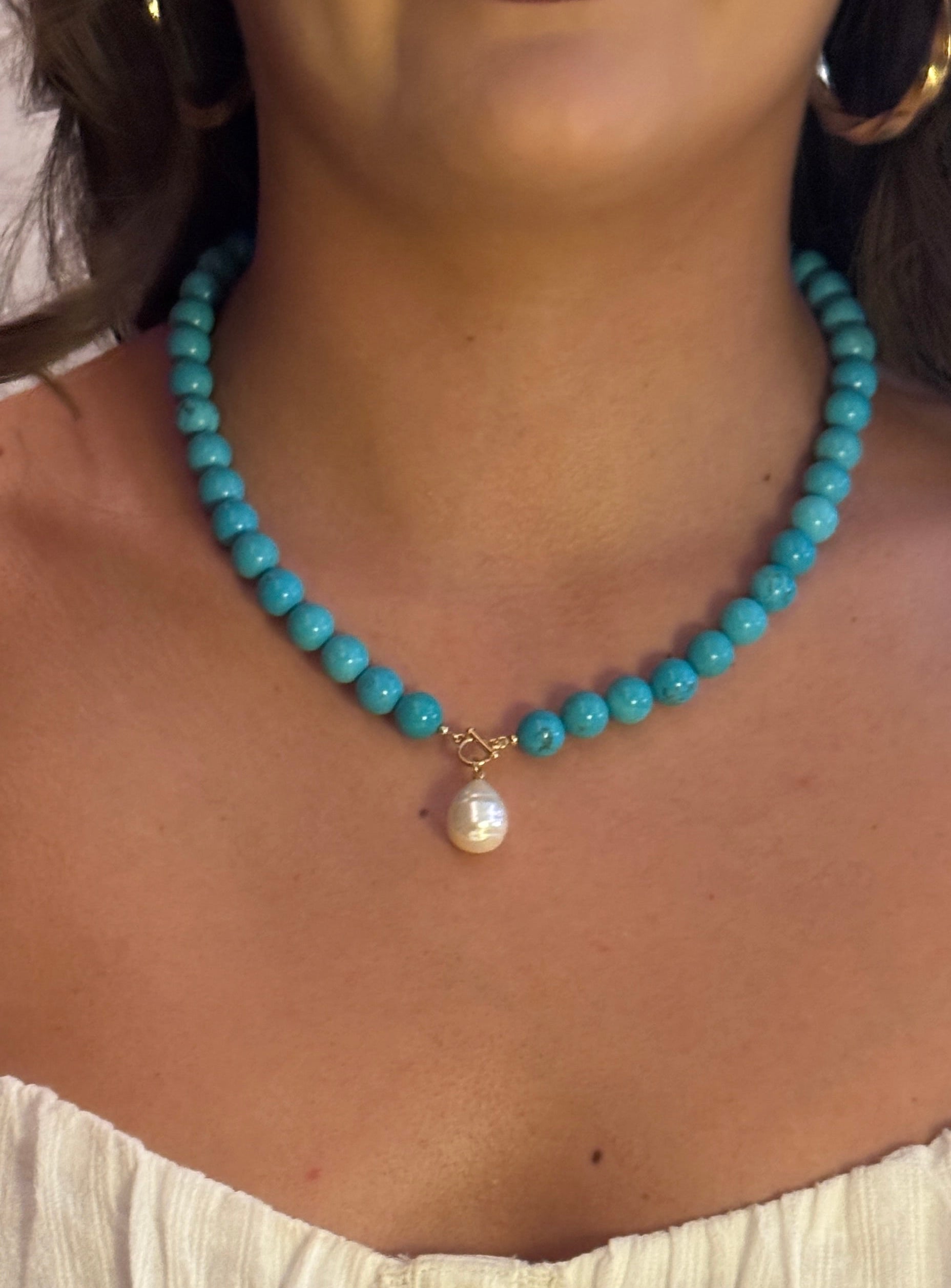 Woman wearing a turquoise beaded necklace with a pearl pendant, holding a glass of red wine.