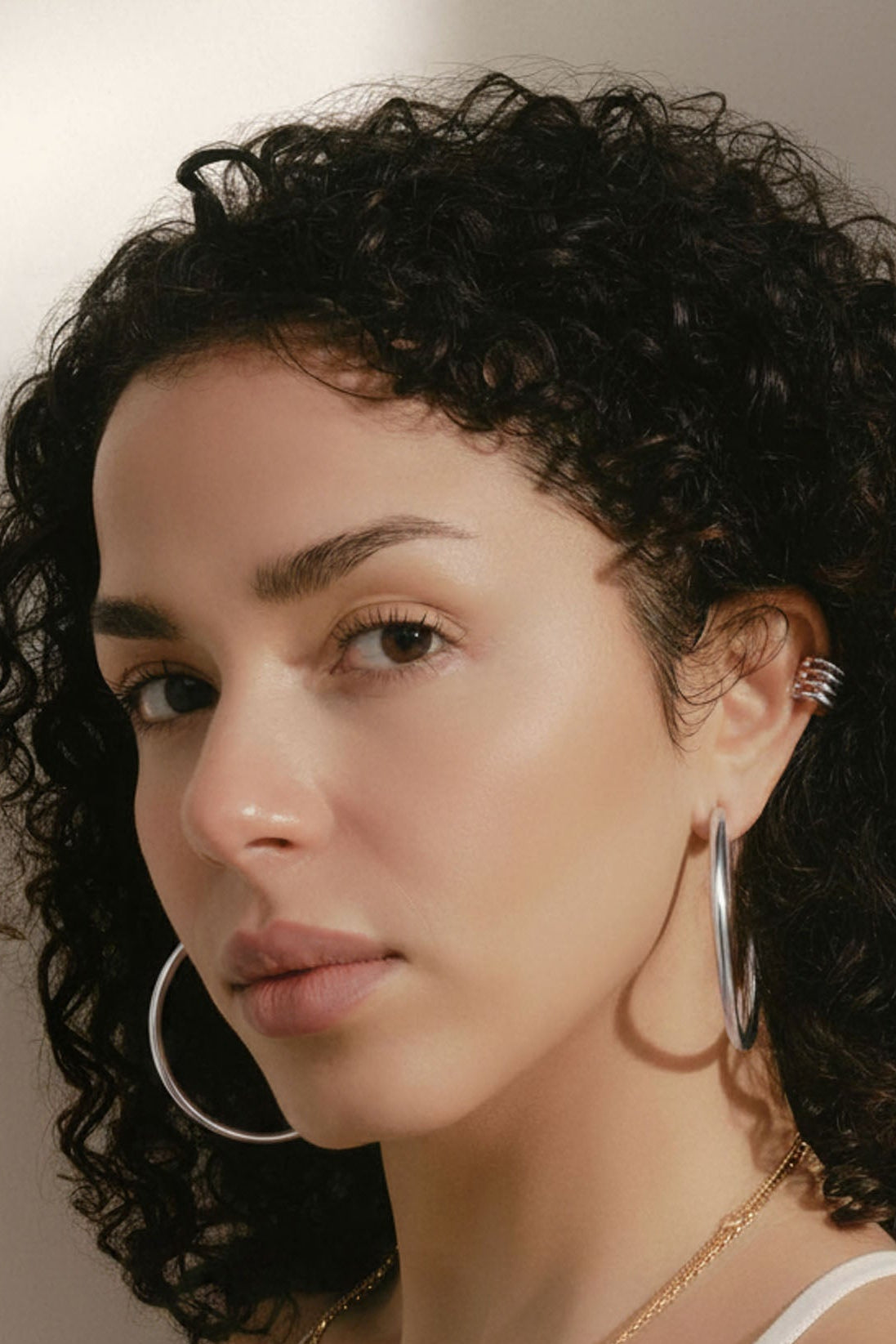 A close-up portrait of a woman with curly dark hair wearing large gold hoop earrings, a delicate gold chain necklace, and a gold ear cuff, paired with a white camisole top against a warm neutral background.