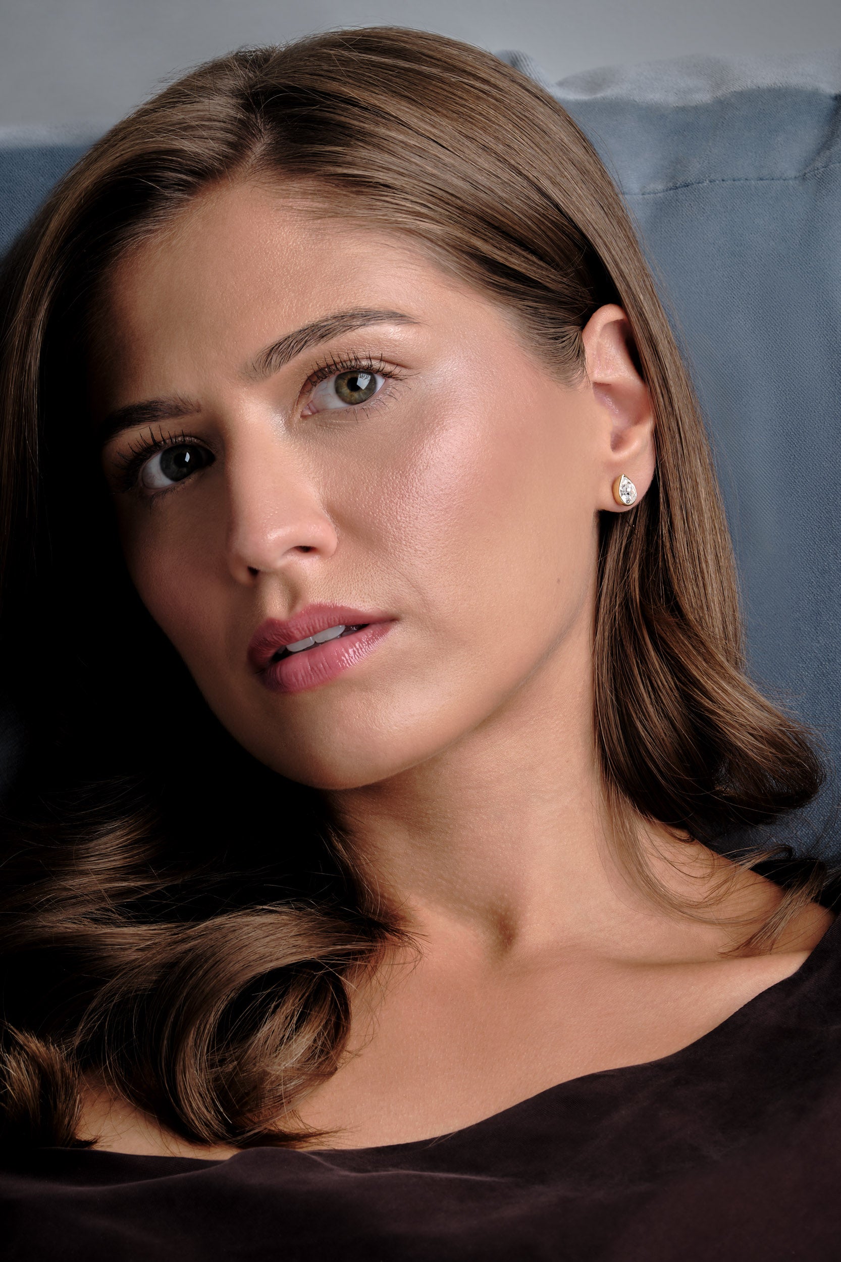 A close-up portrait of a woman with long brown hair, wearing a small stud earring, set against a dark blue-gray background.