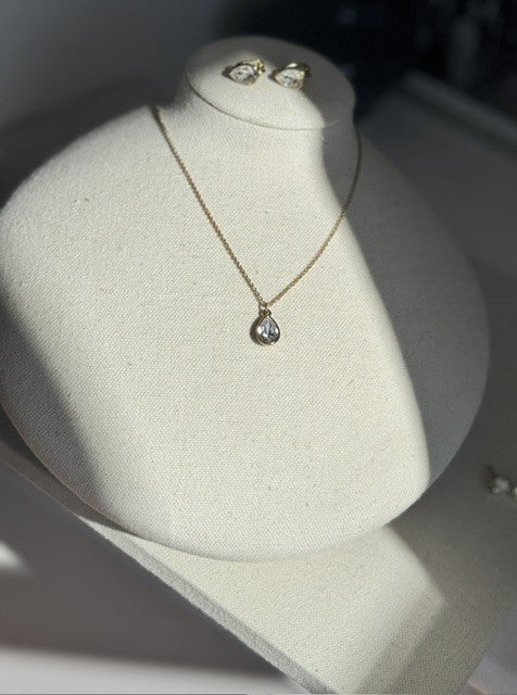A delicate gold necklace with a teardrop-shaped clear gemstone pendant, displayed on a cream-colored jewelry bust. Matching gold stud earrings are partially visible on the top of the bust.