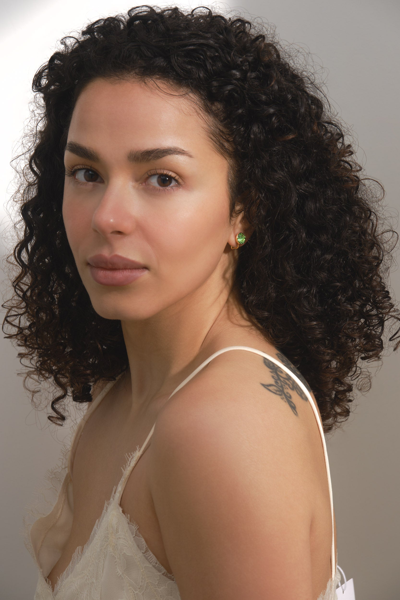 Woman with curly hair wearing a white top against a neutral background and green crystal earrings