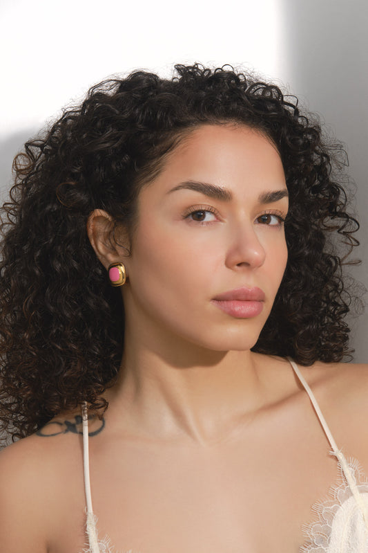 Woman with curly hair wearing a white lace top against a neutral background wearing pink and gold earrings