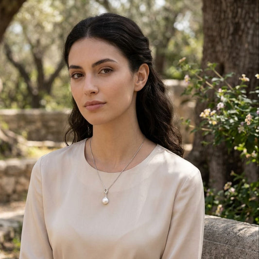 A woman with dark hair tied back sits outdoors wearing a light beige long-sleeve blouse and a delicate silver necklace with a white pearl pendant. The background shows a blurred natural setting with trees and stone.