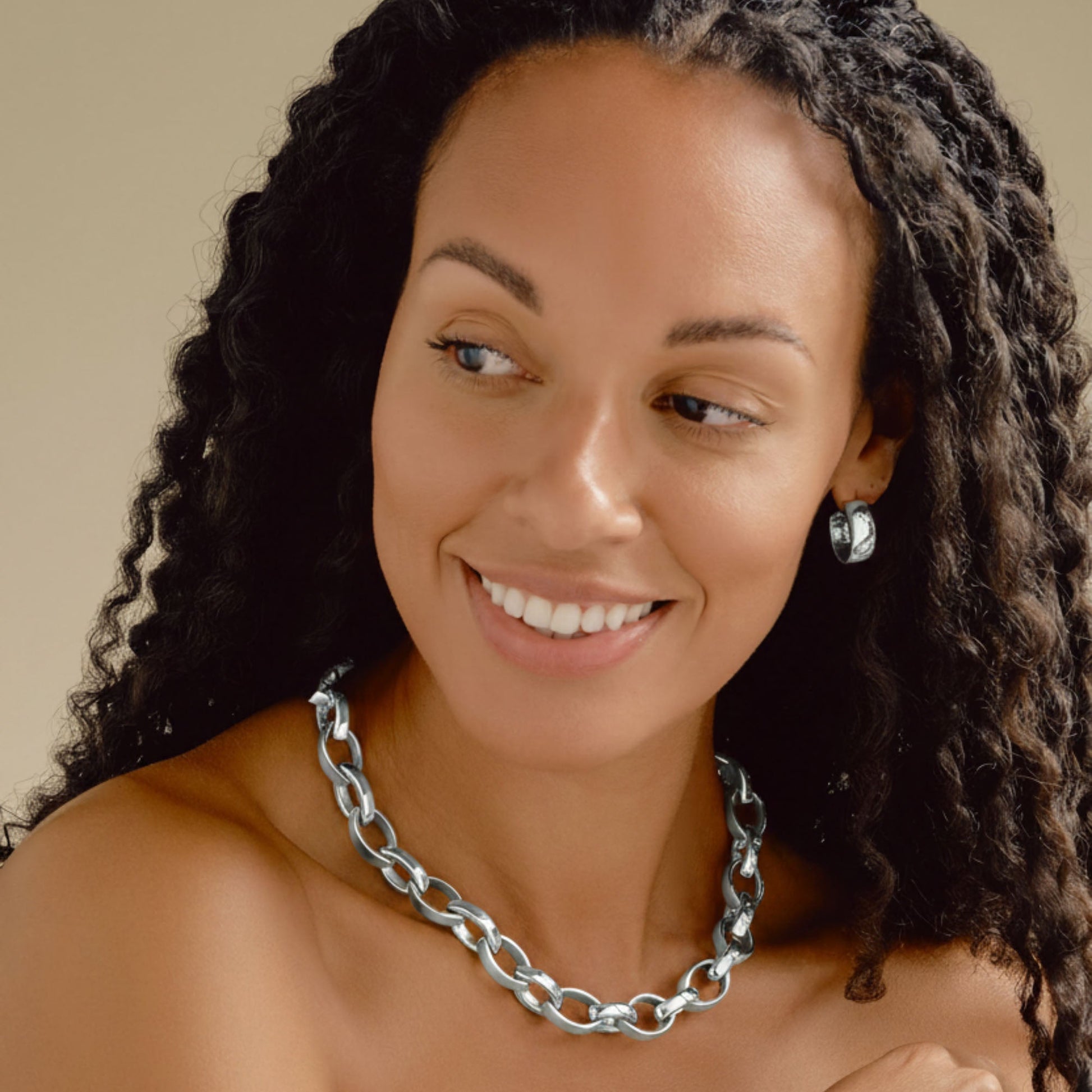 A smiling woman with long curly black hair models a silver chain necklace and matching silver chain bracelet, along with a silver cuff bracelet, against a warm beige background.