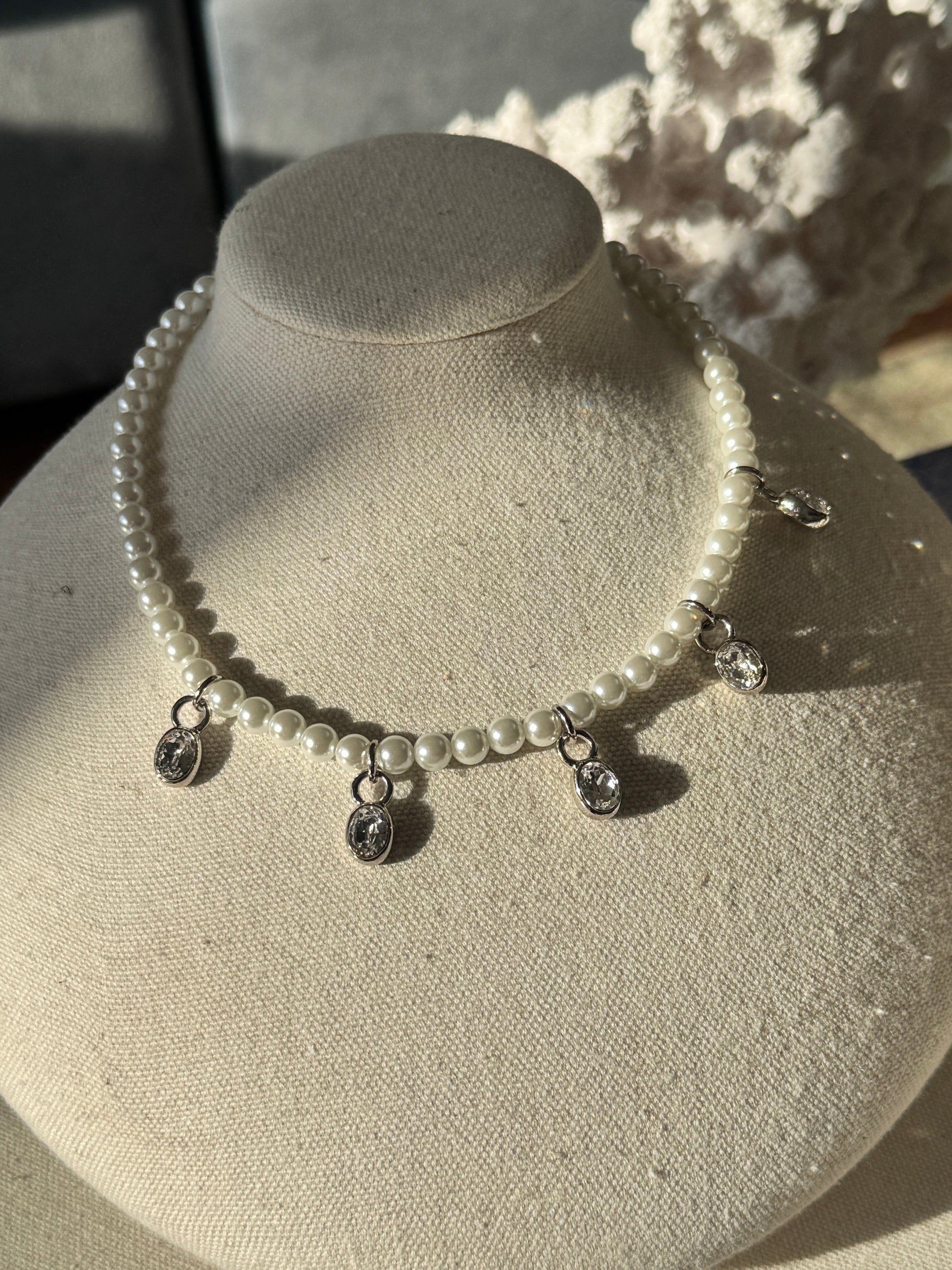 A pearl beaded necklace with silver-toned metal charms and drop pendants, displayed on a cream fabric-covered jewelry bust.