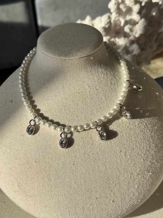 A pearl beaded necklace with silver-toned metal charms and drop pendants, displayed on a cream fabric-covered jewelry bust.