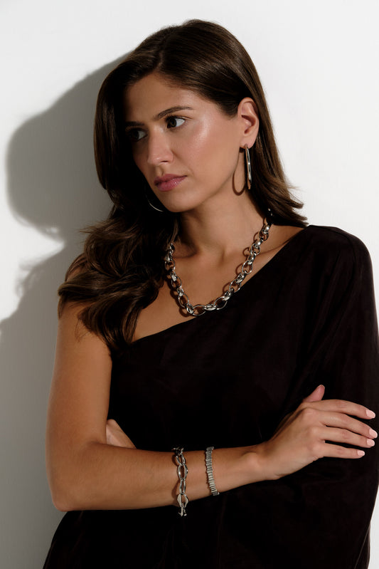 A woman with long brown hair wearing a dark brown one-shoulder top and silver chain jewelry, posing against a white wall with a soft shadow.