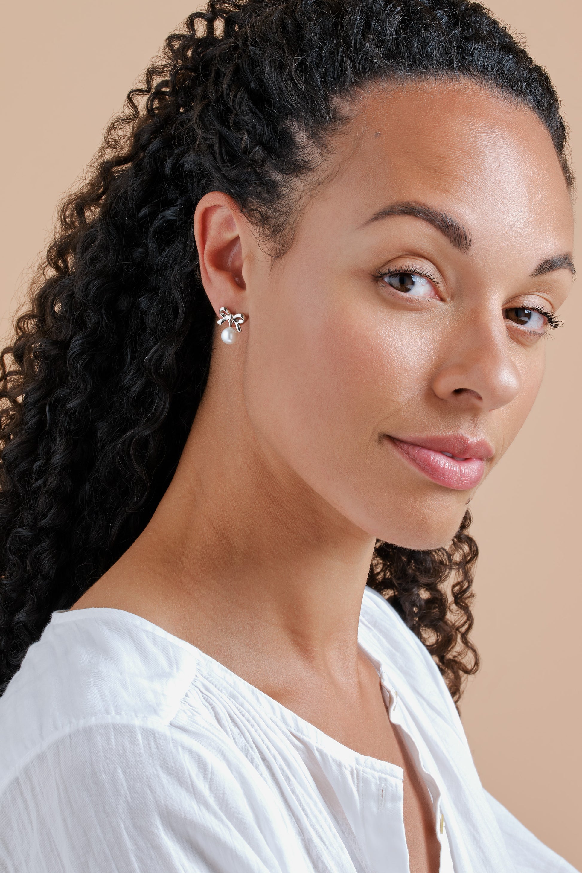 Woman with styled hair and earrings against a beige background wearing platinum bow earrings