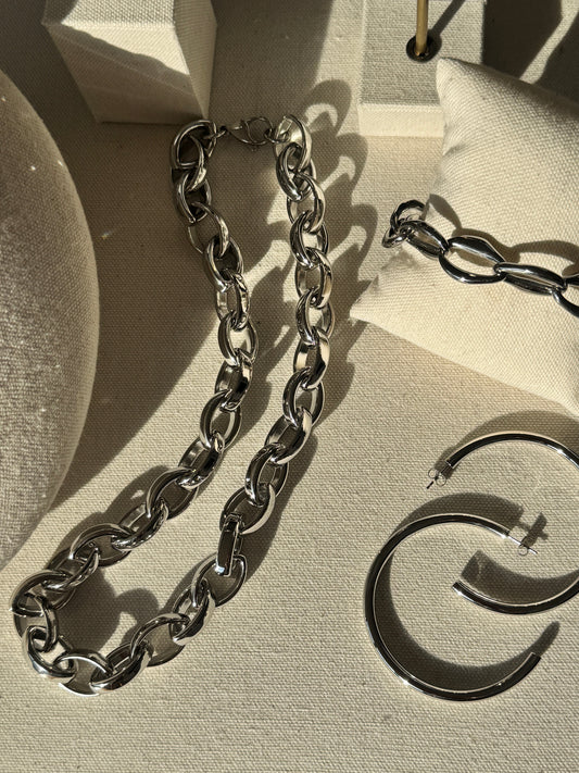 A silver-toned jewelry set displayed on a beige fabric jewelry box, featuring a chunky curb chain necklace, a smaller matching chain segment, and a pair of thick curved hoop earrings.