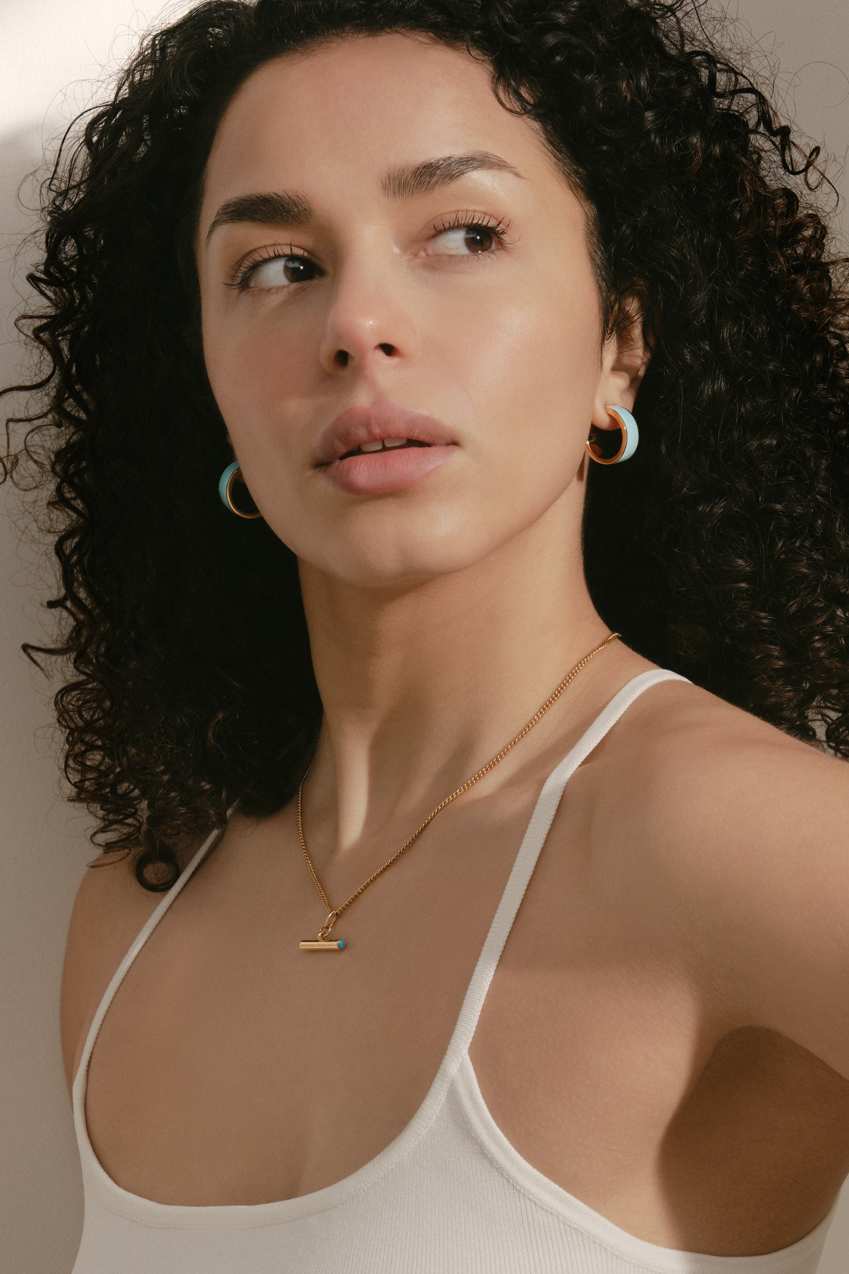 Woman wearing a white tank top and gold necklace with a small pendant.