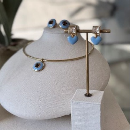 Gold jewelry display set featuring a bangle bracelet, stud earrings, and a dangle necklace with light blue heart and evil eye charms, arranged on a white display bust and stand.