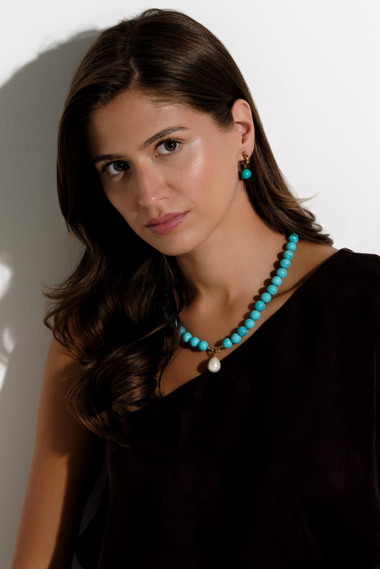 A woman with long brown hair models a turquoise beaded necklace and matching drop earrings against a white wall.